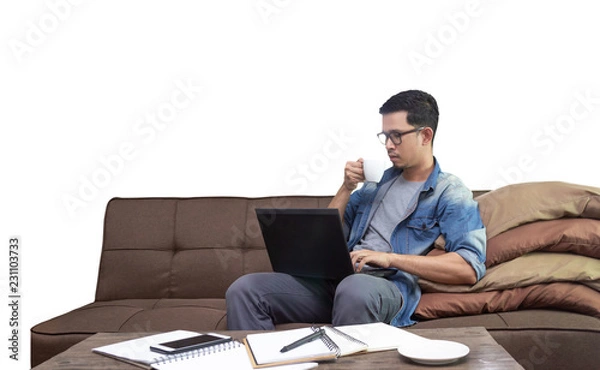 Obraz Asian guy drinking coffee while using laptop sitting on sofa working as freelance at home with clipping path for graphic design