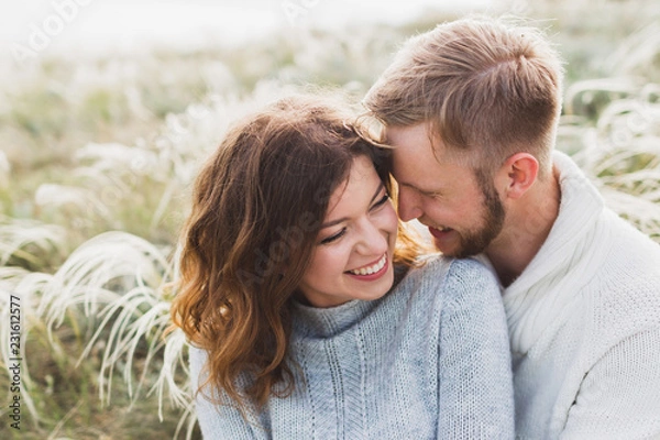 Fototapeta Happy young loving couple sitting in feather grass meadow, laughing and hugging, casual style sweater and jeans