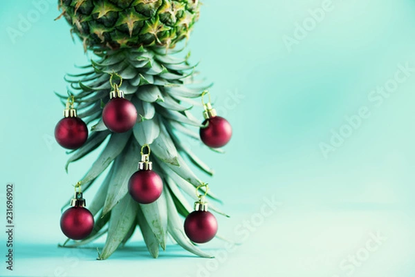 Fototapeta Creative Christmas tree made of pineapple and red bauble on blue background, copy space. Greeting card, decoration for new year party. Holiday concept.