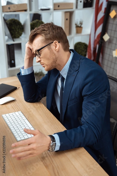 Obraz tired young businessman in eyeglasses gesturing by hand at table with computer keyboard and mouse in office