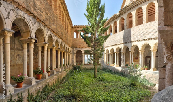 Fototapeta The cloister with romanesque arches, a fir tree and medieval frescos during a sunny day in the Colegiata de Santa Maria la Mayor of Alquezar, Spain