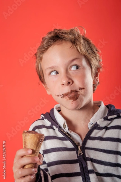 Obraz close up of one young blond and cute boy eating and holding with his hand one chocolate ice-cream over a red background in studio. He has a chocolate stains on his face and a mischievous look.