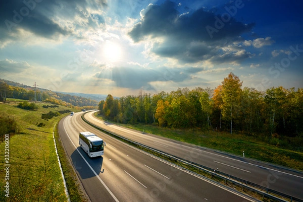 Fototapeta Trzy białe autobusy jeżdżące asfaltową autostradą pomiędzy lasem liściastym w jesiennych barwach pod promieniującym słońcem i dramatycznymi chmurami. Widok z góry.