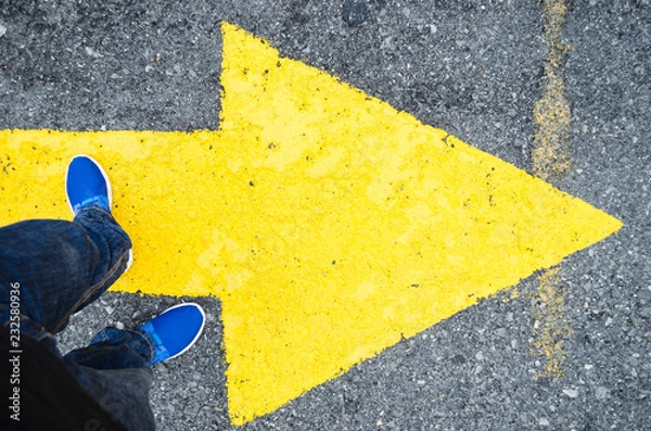 Fototapeta close up on the legs of a man standing on a yellow arrow in the direction to the right, concept of decisions in life.