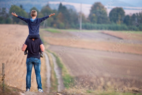 Fototapeta Back view of athletic father carrying on shoulders son walking through grassy field on blurred foggy green trees and blue sky background. Active lifestyle, family relations, weekend activity concept.