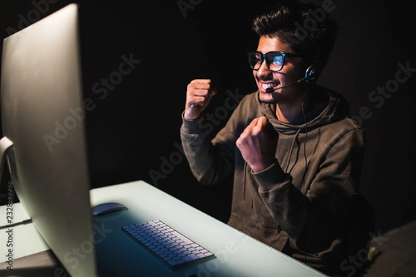 Fototapeta Ecstatic asian gamer boy rejoicing victory while playing video games on computer in dark room wearing headphones and using backlit keyboard