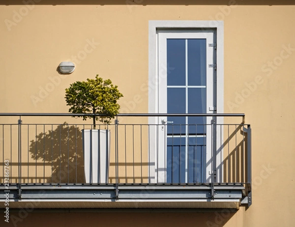 Fototapeta Sunlit beige wall and balcony with bonsai tree and railing casting hard shadows