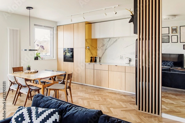 Fototapeta Cozy scandinavian open space with dining, living room and kitchen with design sofa and family table. Bright and sunny room with big windows and brown wooden parquet.	
