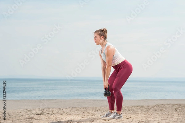 Fototapeta Young attractive woman dressed in pink leggings doing training trying hard in exercises with a kettle bell by the seaside. Fitness woman working out at the beach. Fitness Concept.