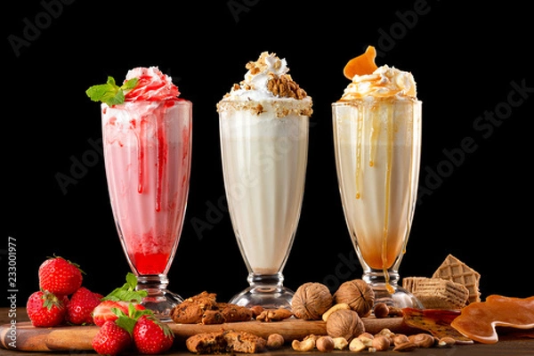 Fototapeta Three sweet milkshakes with nuts, caramel, strawberry and whipped cream at a wooden board on table background.