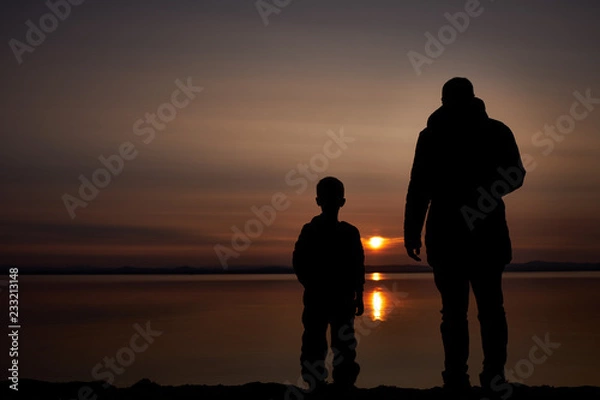 Obraz Silhouettes of the father and his little son stand on the bank of the river and watch the beautiful sunset.