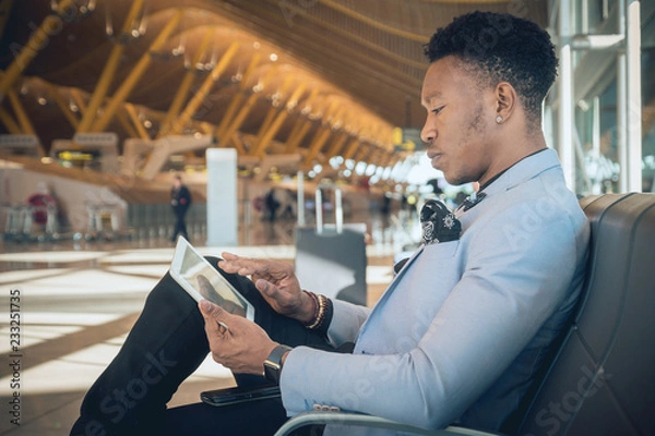 Obraz Young businessman seated in the airport checking a tablet and carrying a suitcase waiting for his flight