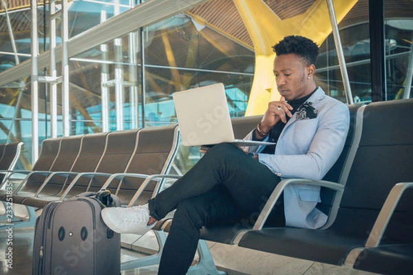 Obraz Young businessman is seated in the airport working with a laptop and carrying a suitcase waiting for his flight
