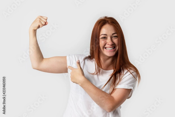 Fototapeta Portrait smiling woman in white t-shirt , isolated on gray background. Showing biceps muscle having lifted arm with clenched fist