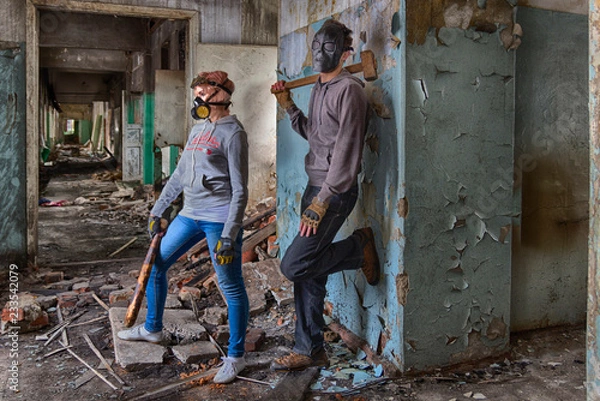 Fototapeta A pair of cyberpunk hooligans: a girl in gas mask and steampunk goggles with a bat and a guy in skull mask with a sledgehammer, standing in the middle of the ruins of an old building. street band