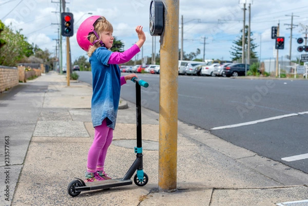 Obraz Young girl in pink outfit and pink helmet riding a kick-scooter pressing button at pedestriam crossing