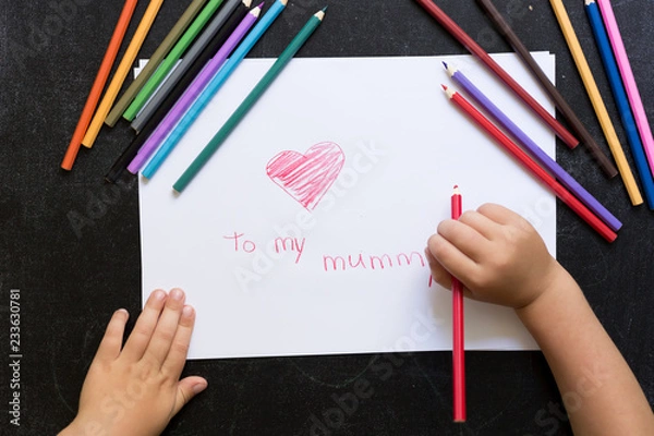 Fototapeta Kid's hands with pencil draws heart on white paper. Mother's day concept. Celebration. Hand made card. Top view. To my mummy words.