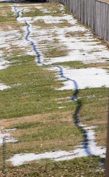 Fototapeta Grass showing through between clumps of melting snow behind a chain link fence.