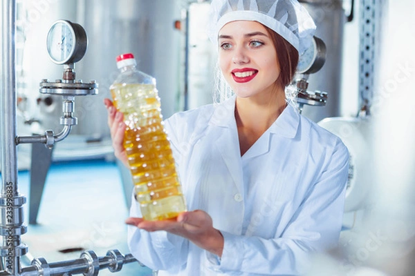 Fototapeta A young girl in white overalls is holding a bottle of sunflower oil in her hands against the background of the factory equipment of the food industry