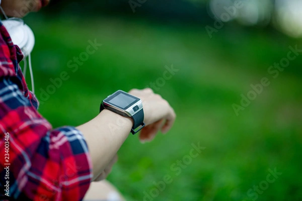 Obraz The girl watches the watch in hand, watches the time in a black watch, wears a red shirt and a green background. And there is a copy space.
