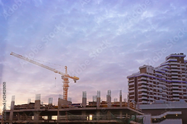 Fototapeta construction of high-rise buildings with yellow tower crane at sunset and dark blue clouds