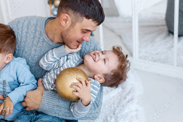 Fototapeta A daddy with his two sons twins in front of the christmas tree  with candles and gifts hugging and having fun. New year's eve. Cozy holiday at the fir-tree. love, happiness and big family concept