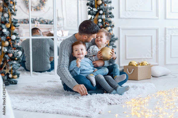 Obraz A a daddy with his two sons twins in front of the christmas tree  with candles and gifts hugging and having fun. New year's eve. Cozy holiday at the fir-tree. love, happiness and big family concept