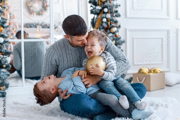 Obraz A a daddy with his two sons twins in front of the christmas tree  with candles and gifts hugging and having fun. New year's eve. Cozy holiday at the fir-tree. love, happiness and big family concept