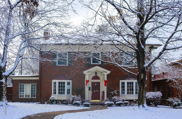 Fototapeta Beautiful brick house with bay windows with Christmas tree showing through and decorated pillars and sled on porch in snow framed by winter trees