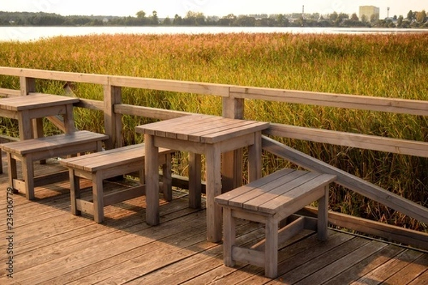 Fototapeta Wooden tables and benches on the lake with reeds waiting for lovers to watch the sunset.