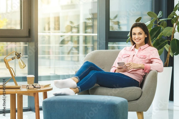 Fototapeta Searching more information. Beautiful young smiling woman looking at her smart phone while sitting in a big comfortable chair at home.