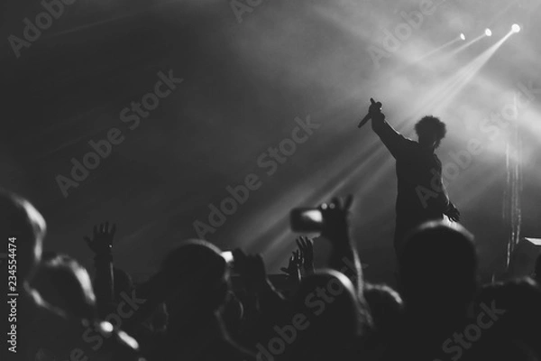 Obraz Black and white photo of frontman silhouette in a stage backlights with hand with microphone raised up to the crowd of people