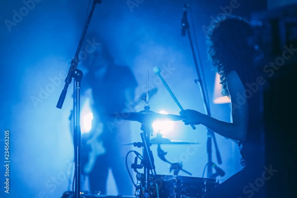 Obraz  Female drummer with long curly hair in front of singing guitarist on a stage in a bright blue lights playing rock show