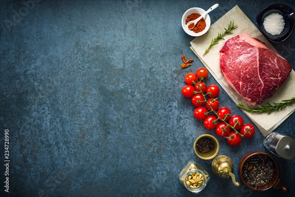 Obraz Raw marbled beef steak on dark concrete background with salt, herbs and tomatoes. Top view and copyspace.