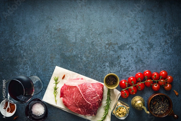Obraz Raw marbled beef steak on dark concrete background with salt and herbs, tomatoes and glass of red wine. Top view and copyspace