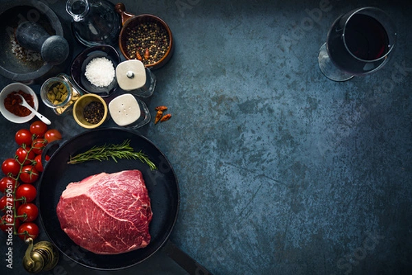 Obraz Raw marbled beef steak on cast iron pan with salt and herbs, tomatoes and glass of red wine. Top view and copyspace.