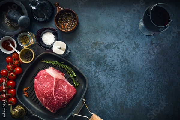 Obraz Raw marbled beef steak on a grill pan with salt, herbs and tomatoes and glass of red wine. Top view and copyspace.