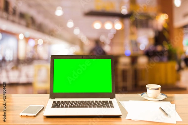Fototapeta Mockup image of laptop with blank green screen on wooden table blurred images In the coffee shop at shopping mall background.