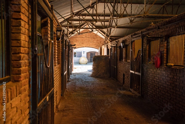 Fototapeta Inside a bricks stable looking trough a door  two hay rolls and horses stals