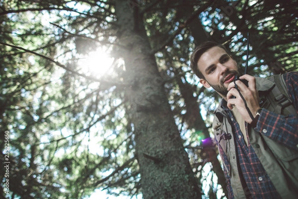 Fototapeta Concept of experienced journey and connection. Low angle portrait of cheerful bearded man talking on portable radio set while walking in green sunny forest