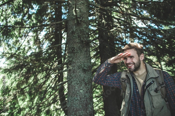 Fototapeta Concept of interesting journey and adventure. Waist up portrait of smiling young male putting his hand on forehead and trying to see something among green trees