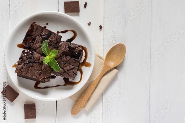 Obraz A plate of brownies with chocolate sauce on white wooden background, mint leaf on top, top view