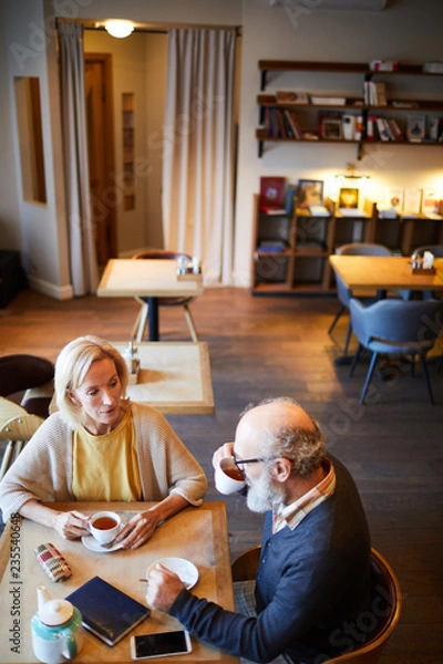 Fototapeta Senior man drinking tea while sitting by table in front of his colleague during work planning
