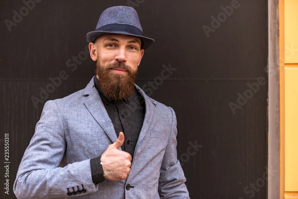 Fototapeta stylish man with a beard in a hat on the street showing hand signs