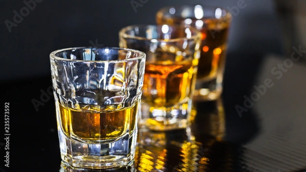 Obraz Shots de whisky en fila sobre fondo negro y luz directa en ellos