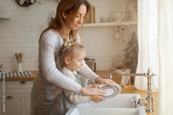 Fototapeta A small child with interest helps her mother with household chores. The girl is happy to learn to wash dishes