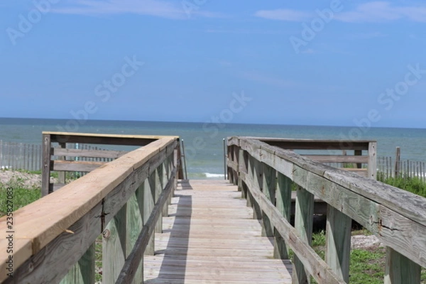 Fototapeta Perspective photograph of wood boardwalk railing beach access horizon blue sky, turquoise ocean and green grass.