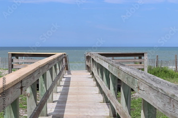 Fototapeta Perspective photograph of wood boardwalk railing beach access horizon blue sky, turquoise ocean and green grass.
