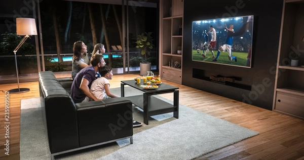 Obraz A family is watching a soccer moment on the TV and celebrating a goal, sitting on the couch in the living room.