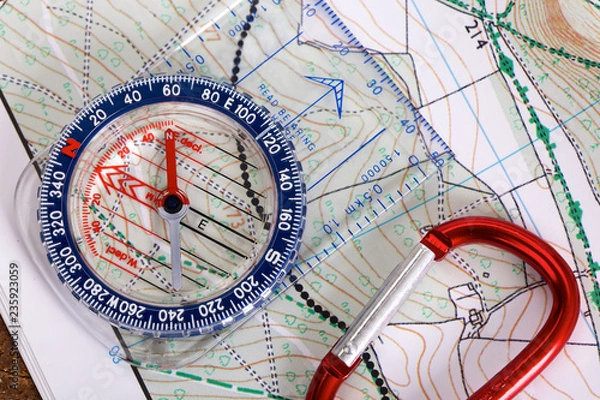 Fototapeta Close up of a modern plastic compass with scales and rulers, wooden background and detailed hiking map
